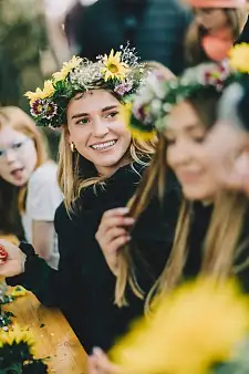 Das Bild zeigt eine fröhliche junge Frau mit einem Blumenkranz, die an einem Holztisch mit anderen Menschen sitzt und sich unterhält.