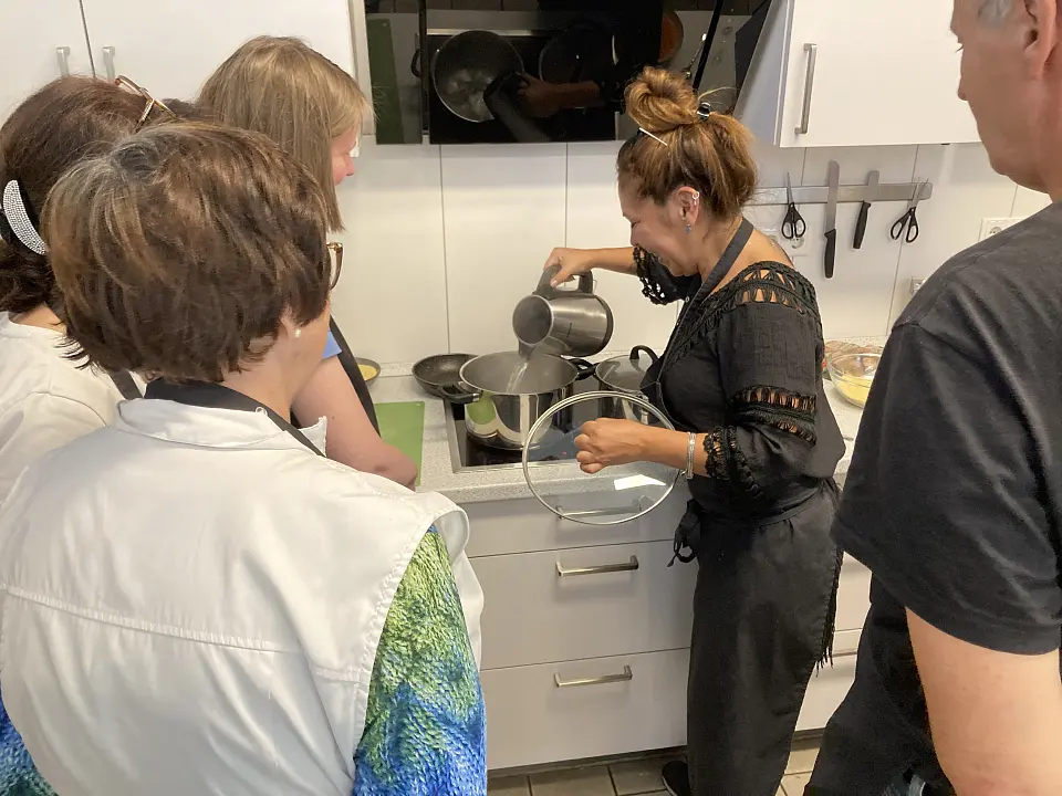 Das Bild zeigt vier Personen, die um einen Herd herumstehen an dem eine Person Wasser in einen Topf gie&szlig;t.