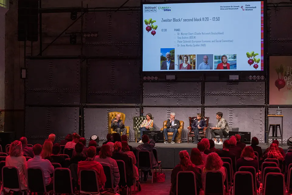 Bild einer B&uuml;hnensituation mit 4 Personen. Die Personen sind von links nach rechts: Peter Schmidt (Vorsitzender der Fachgruppe Landwirtschaft, l&auml;ndliche Entwicklung und Umwelt im Europ&auml;ischen Wirtschafts- und Sozialausschuss), Tina Andres (Vorstandsvorsitzende Der Bund &Ouml;kologische Lebensmittelwirtschaft), Dr. Werner Ebert (Gesch&auml;ftsf&uuml;hrer Bio St&auml;dte Netzwerk Deutschland), Dr. Anne Monika Spallek (Mitglied des Bundestages , Ausschuss f&uuml;r Ern&auml;hrung und Landwirtschaft), Dan Anthes (Moderator der Veranstaltung). Es handelt sich um eine Diskussionsrunde.