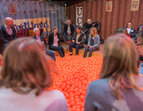 Mehrere Personen sitzen in einem Kreis mit ihren Füßen in einem Bällebad mit orangen Bällen und hören einer Person zu. 
˜ Bildnachweis: Christian Arne de Groot