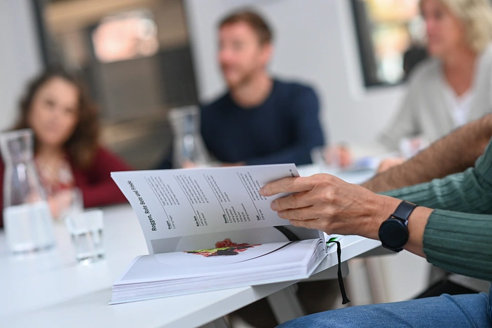 Fortbildung: Eine Person bl&auml;ttert in einem Heft, w&auml;hrend im Hintergrund andere an einem Tisch sitzen.