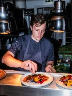 Das Foto zeigt Joscha Frölich, der konzentriert ein Gericht auf einem Teller anrichtet. Auf dem Teller vor ihm befindet sich ein kunstvoll angerichtetes Gericht, das mit verschiedenen Obstsorten (wie Beeren und Pflaumen) und Saucen dekoriert ist. 
&tilde; Bildnachweis: Joscha Frölich