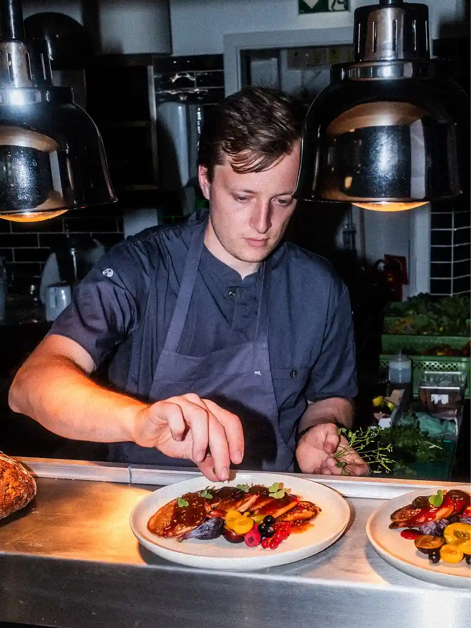 Das Foto zeigt Joscha Fr&ouml;lich, der konzentriert ein Gericht auf einem Teller anrichtet. Auf dem Teller vor ihm befindet sich ein kunstvoll angerichtetes Gericht, das mit verschiedenen Obstsorten (wie Beeren und Pflaumen) und Saucen dekoriert ist.