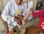 Drei Kinderhände greifen nach Hafer aus einem Glas. Ein Kind füllt die Haferkörner in eine manuelle Haferquetsche. In der darunterstehenden Schüssel und in der Auslassrinne liegt bereits gequetschter Hafer. 
˜ Bildnachweis: Verein Sozialökologie e.V.