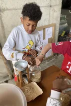Drei Kinderhände greifen nach Hafer aus einem Glas. Ein Kind füllt die Haferkörner in eine manuelle Haferquetsche. In der darunterstehenden Schüssel und in der Auslassrinne liegt bereits gequetschter Hafer. 
&tilde; Bildnachweis: Verein Sozialökologie e.V.