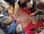 Rund ein Dutzend Kinder und ein paar Erwachsene stehen dicht gedrängt um einen Tisch, auf dem gebackene Brötchen liegen. Die Brötchen sind auf Backpapier verteilt und unterschiedlich bestreut und geformt. Einige Kinder schauen gespannt oder zeigen auf bestimmte Brötchen. Das Bild wurde von oben aufgenommen und vermittelt eine aktive, gemeinschaftliche Stimmung. 
˜ Bildnachweis: Verein Sozialökologie e.V.